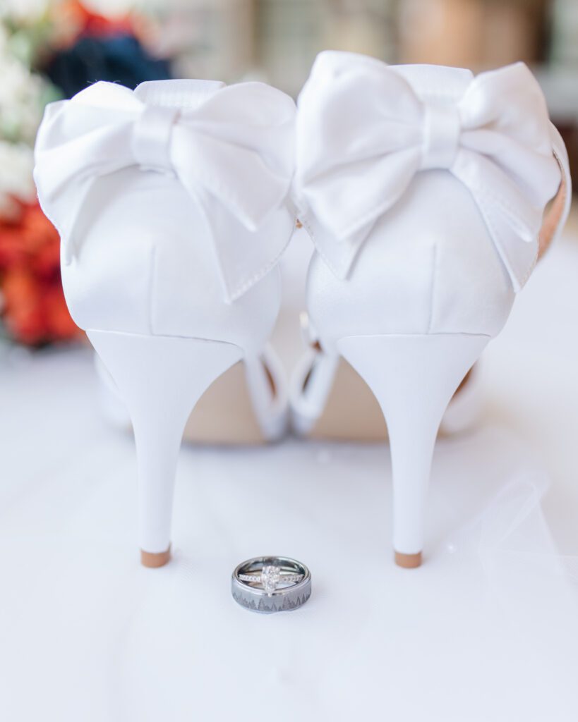 Bridal details of white heals with bows on the backs, rings and flowers in the background