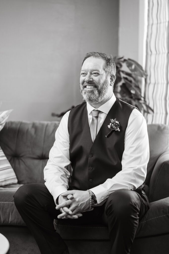 Groom portrait as he sits on couch, smiling at his groomsmen