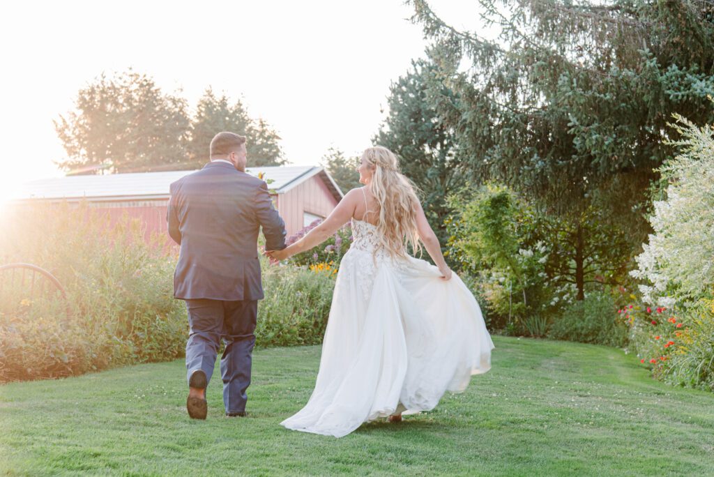 Bride and Groom holding hands at Washington venue as they walk into the sunset in a field with a red barn at the wedding venue: Lucy's Garden in Ridgefield, WA