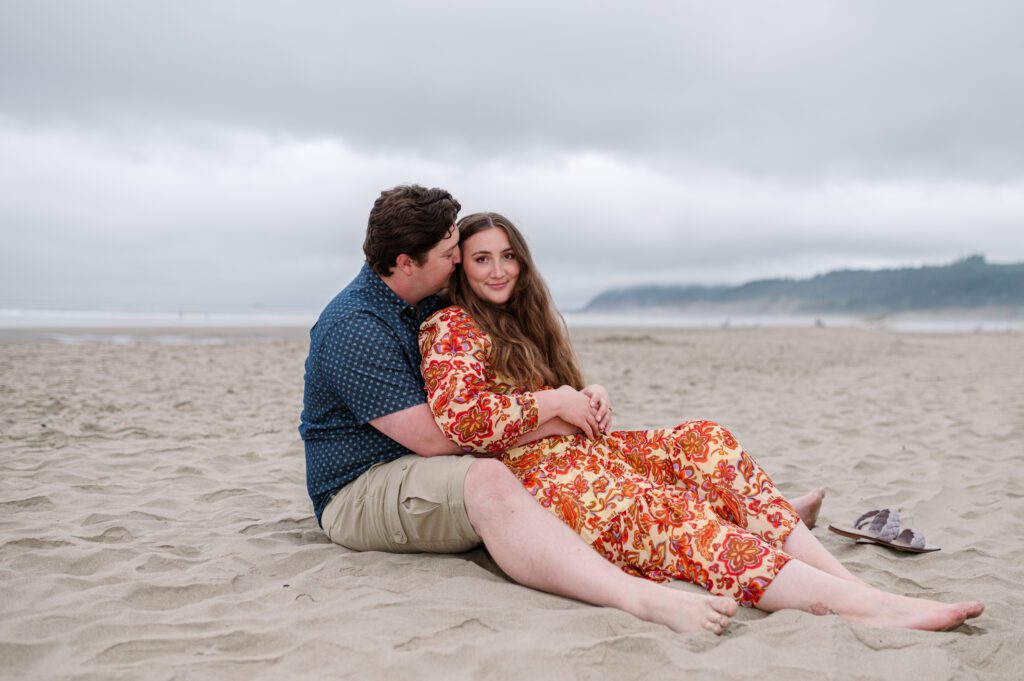 Bride leaning into groom while sitting down at Cannon Beach in Oregon for their engagement session