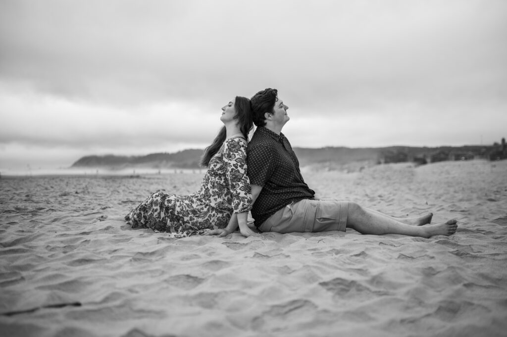 Black and white image of a couple sitting down back to back on the beach at cannon beach in Oregon