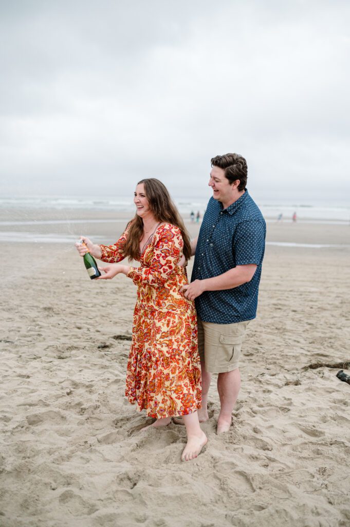 Couple popping champagne on the beach in Cannon Beach, OR while celebrating their engagement