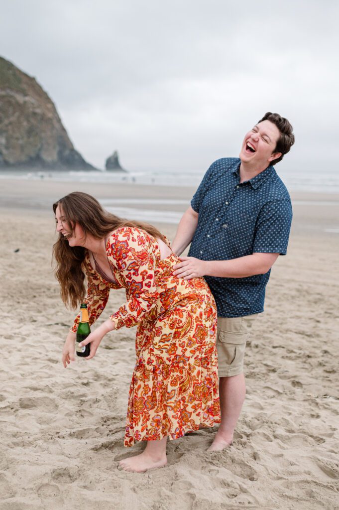 Couple laughing as they pop champagne at cannon beach in front of haystack rock for their engagement session in oregon