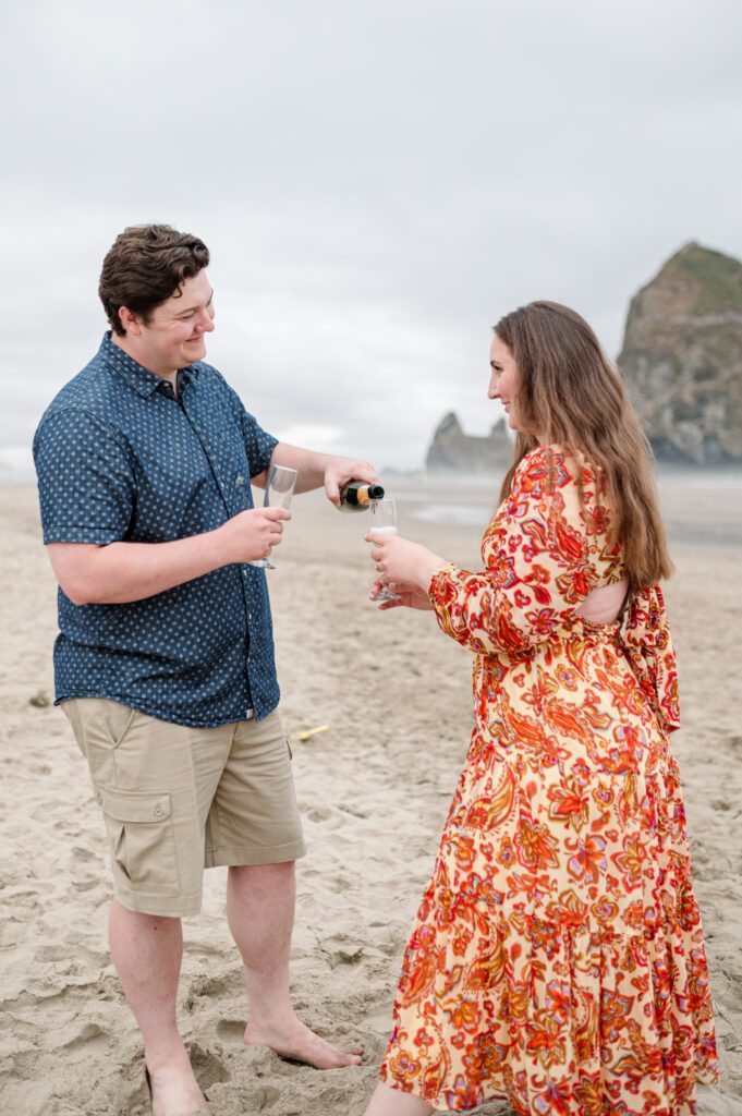 Couple sharing champagne in front of haystack rock in cannon beach oregon for their engagement session on the oregon coast