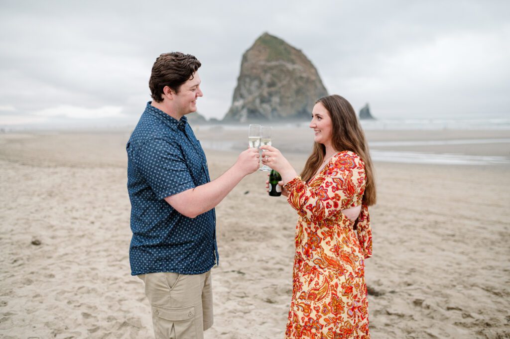 couple saying cheers with their champagne glasses on the oregon coast for their engagement session in cannon beach