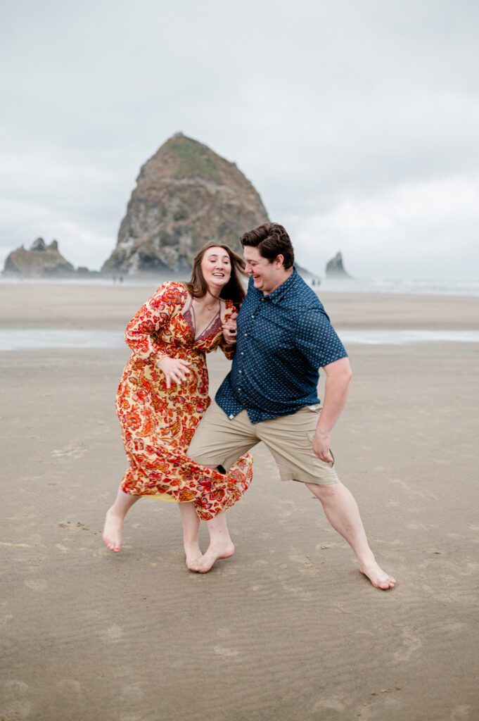 couple walking along the oregon coast in the water and bumping hips and laughing at their engagement session in cannon beach oregon