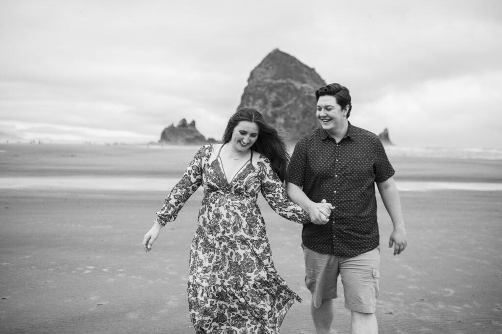 Black and white image of couple holding hands and walking along the oregon coast during their engagement session in cannon beach