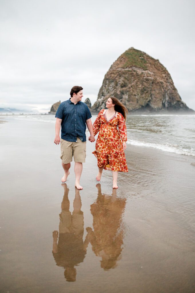 couple holding hands and walking along the oregon coast with their reflection in the water in front of haystack rock in cannon beach oregon for their engagement session