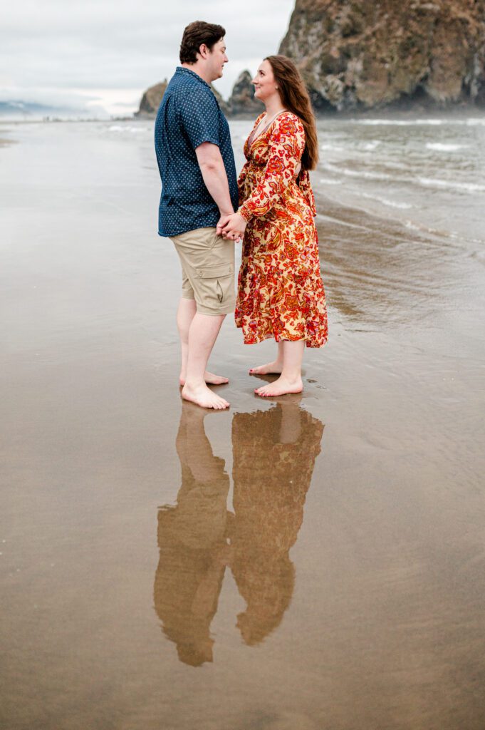 Couple holding hands and looking at each other with their reflection in the wet sand while standing in front of haystack rock at cannon beach oregon for their engagement session