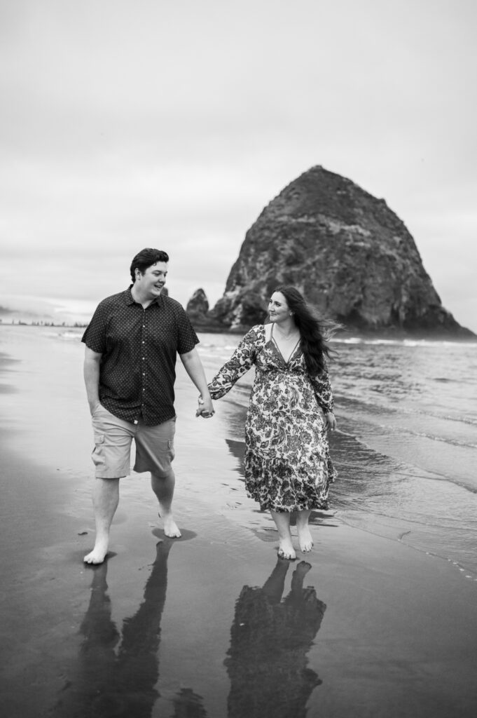 black and white image of an engaged couple holding hands and walking along oreogn coast in front of haystack rock during their engagement session at cannon beach oregon