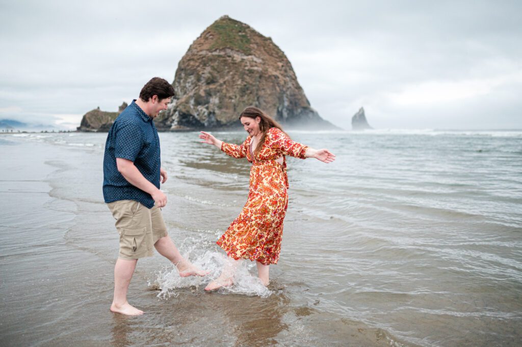 couple kicking water at each other at the oregon coast in front of haystack rock for their engagement session in cannon beach oregon